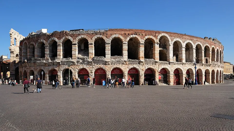 The curved wall of a huge oval amphitheatre in rusticated pinkish stone, two storeys of round arches with red-painted doors at ground level, rising over a wide paved square with people walking across it; a short, taller fragment of wall stands at the left end.