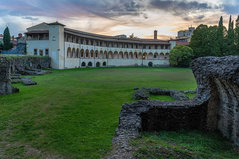 A broad lawn filling the oval of the arena, edged by low broken walls of rubble masonry, with a long two-storey building with arcaded loggias following the curve of the far side under an evening sky.