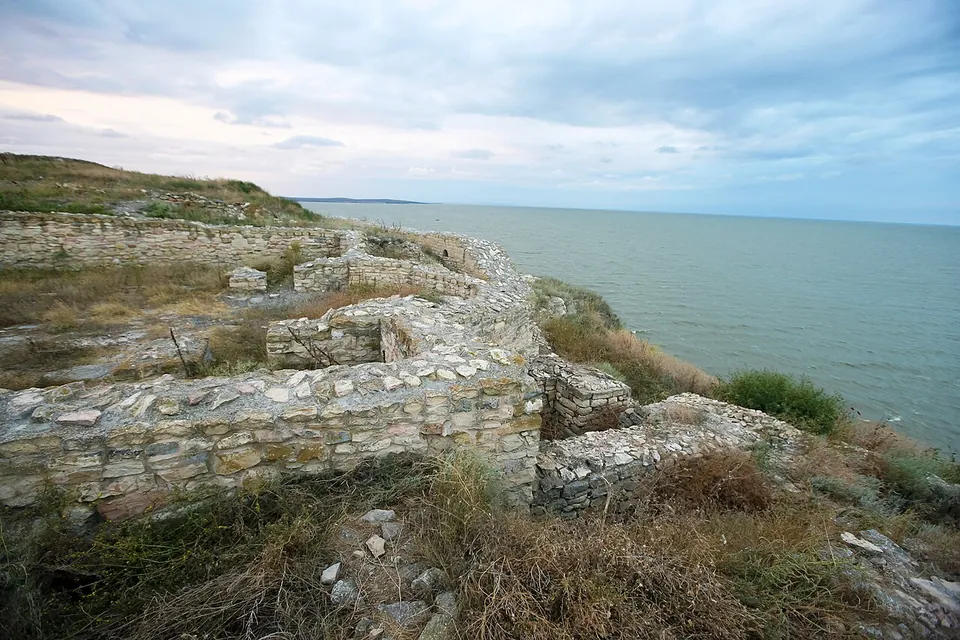 Ruined walls of pale stone running along the grassy edge of a cliff, with the calm grey water of the lake stretching to the horizon under a cloudy sky.