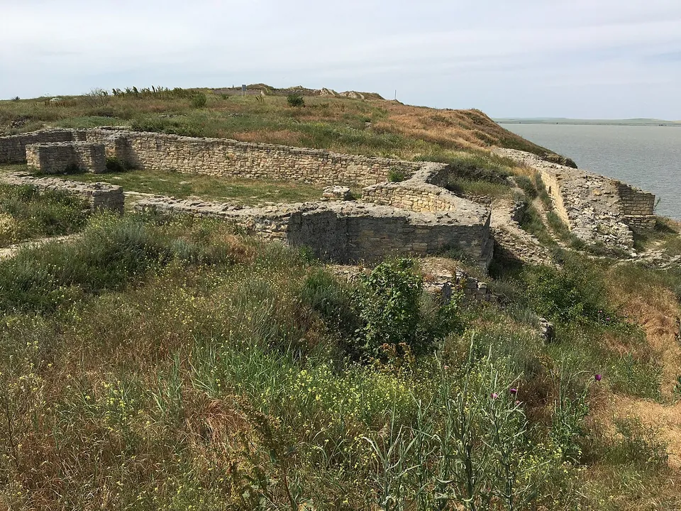 The excavated stone walls of a large building, about head high in places, among long grass and wild flowers on the headland, with the lake beyond.