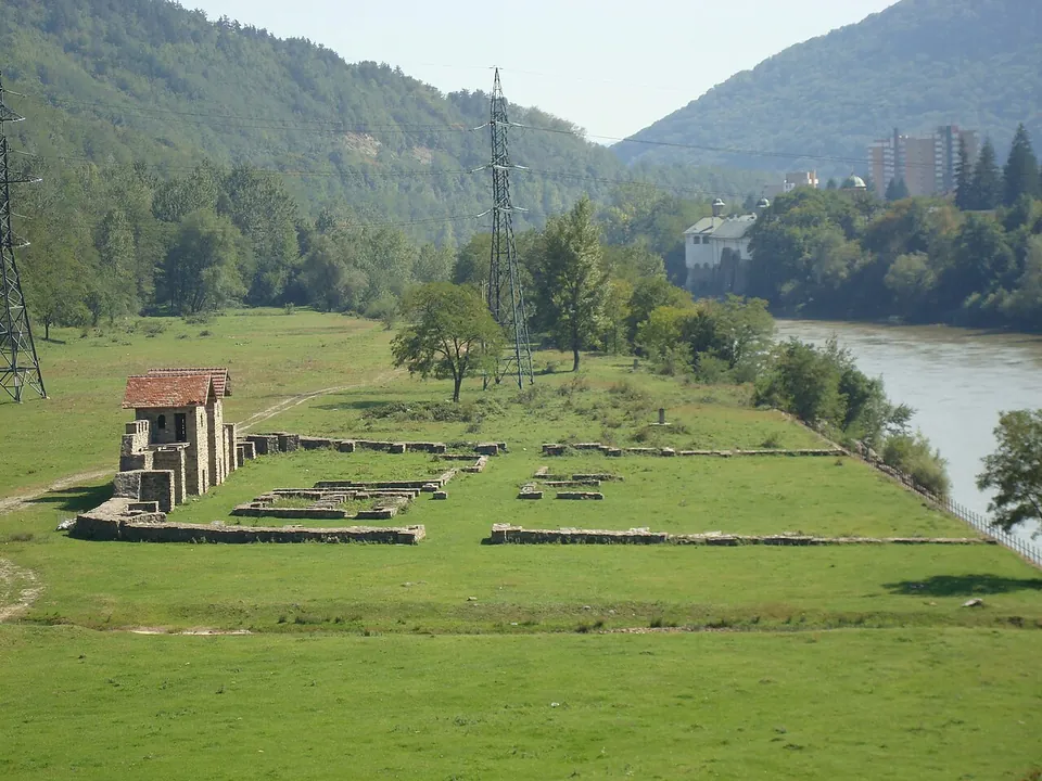A small Roman fort on a flat green terrace beside a wide river, its low foundation walls laid out in the grass around a reconstructed stone gate with tiled roofs, with wooded hills, power lines and a tower block beyond.
