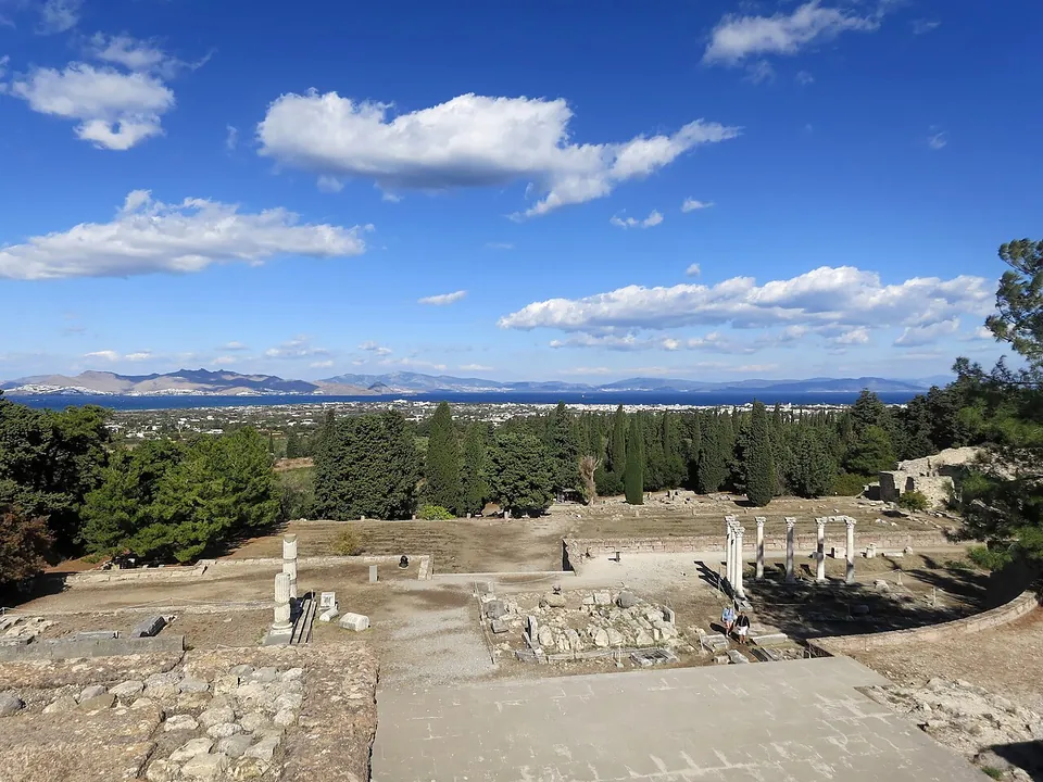 The view from the top of the sanctuary over its lower terraces, stone foundations, steps and a few standing columns, down across a belt of cypresses to the town, the sea and a mountainous coast on the far side.