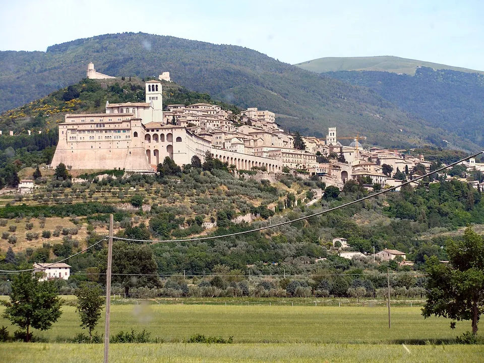 A hill town seen across flat green fields, a long line of pale stone buildings stretched along the slope with a large arcaded building at its left end, a small castle on the bare hilltop above and wooded mountains behind.