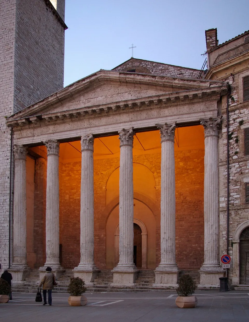 The front of the temple at dusk, six tall fluted columns with leafy capitals carrying a plain triangular pediment, the wall behind them lit orange, with people crossing the paved square in front and a stone tower rising at the left edge.