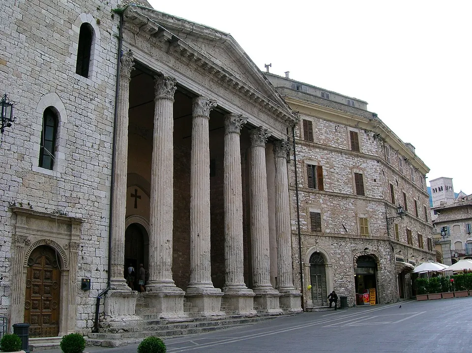 The temple portico seen at an angle from the street, its columns standing on tall square pedestals between the steps, with older stone houses continuing the same frontage on both sides.