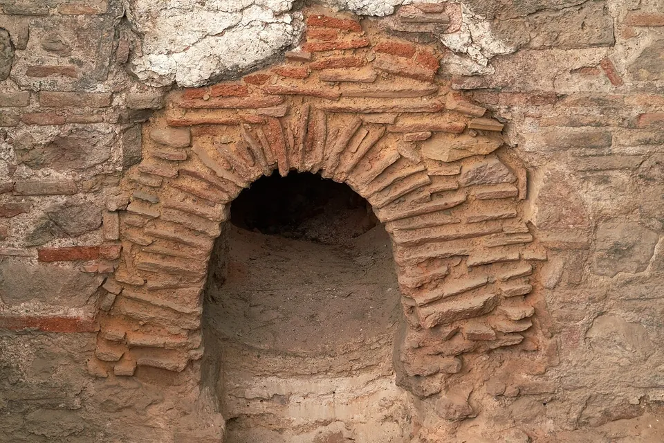 A small round-headed arch of thin red bricks set in a wall of stone and mortar, opening into a dark chamber.