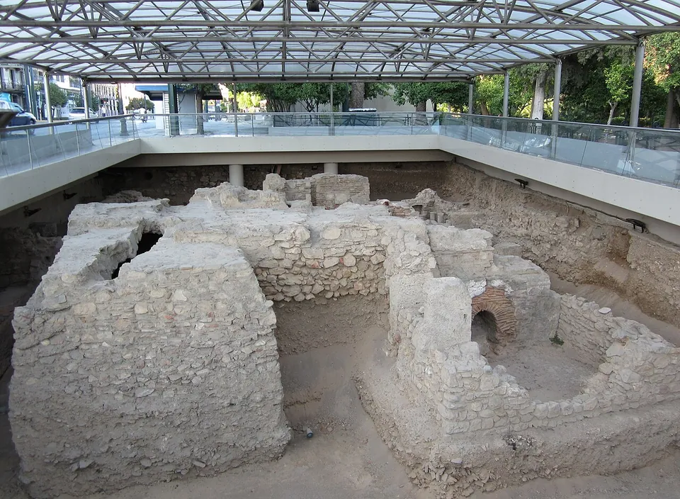 Excavated walls of rubble and squared stone, with a small brick arch, in a pit below street level, under a modern glass and steel roof edged by a railing.