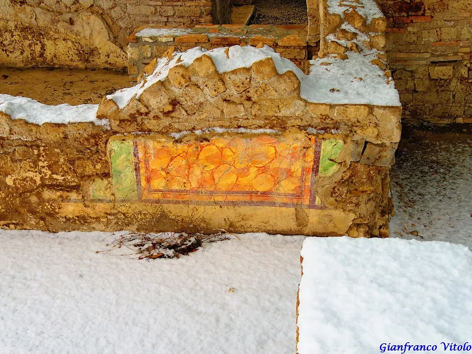 Low walls of excavated rooms, edged with snow and partly plastered in red and ochre, under a spreading metal roof, with a wooden fence in the foreground.