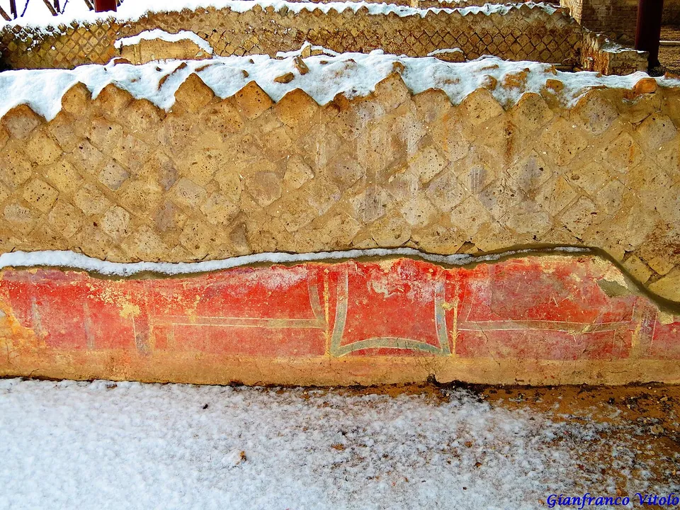 A wall faced with small stones set in a diagonal net pattern, above a band of red-painted plaster divided into panels by thin lines.