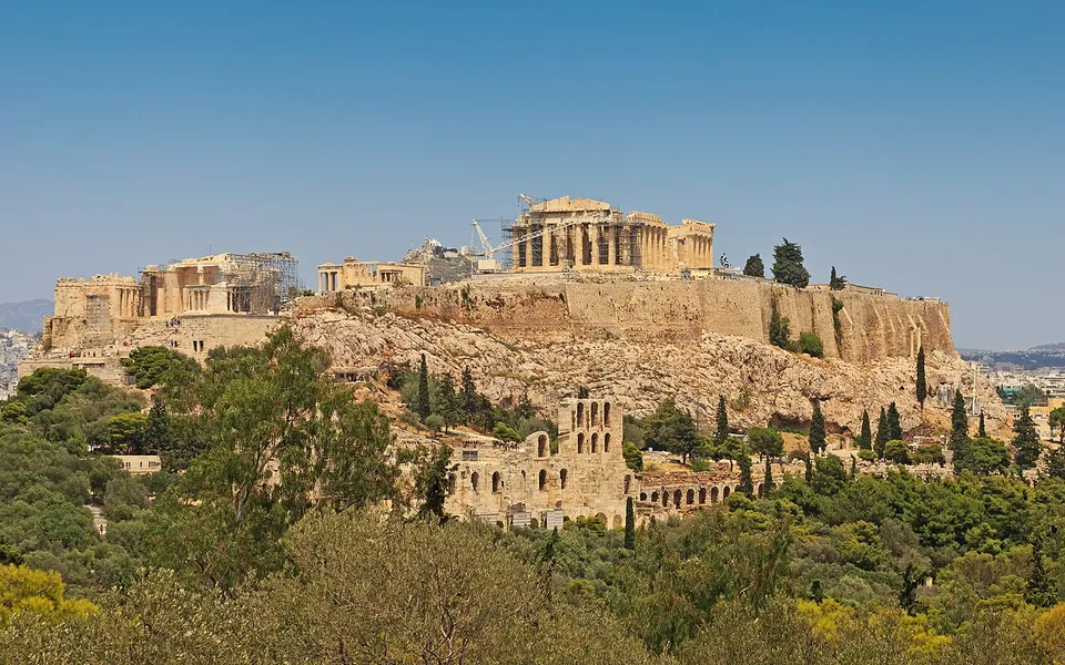 The rock of the Acropolis seen from a wooded hill to the south-west, its long fortification wall and ruined temples along the top under a clear sky, and below the rock a ruined stone facade with tiers of round-headed arches set among pines.