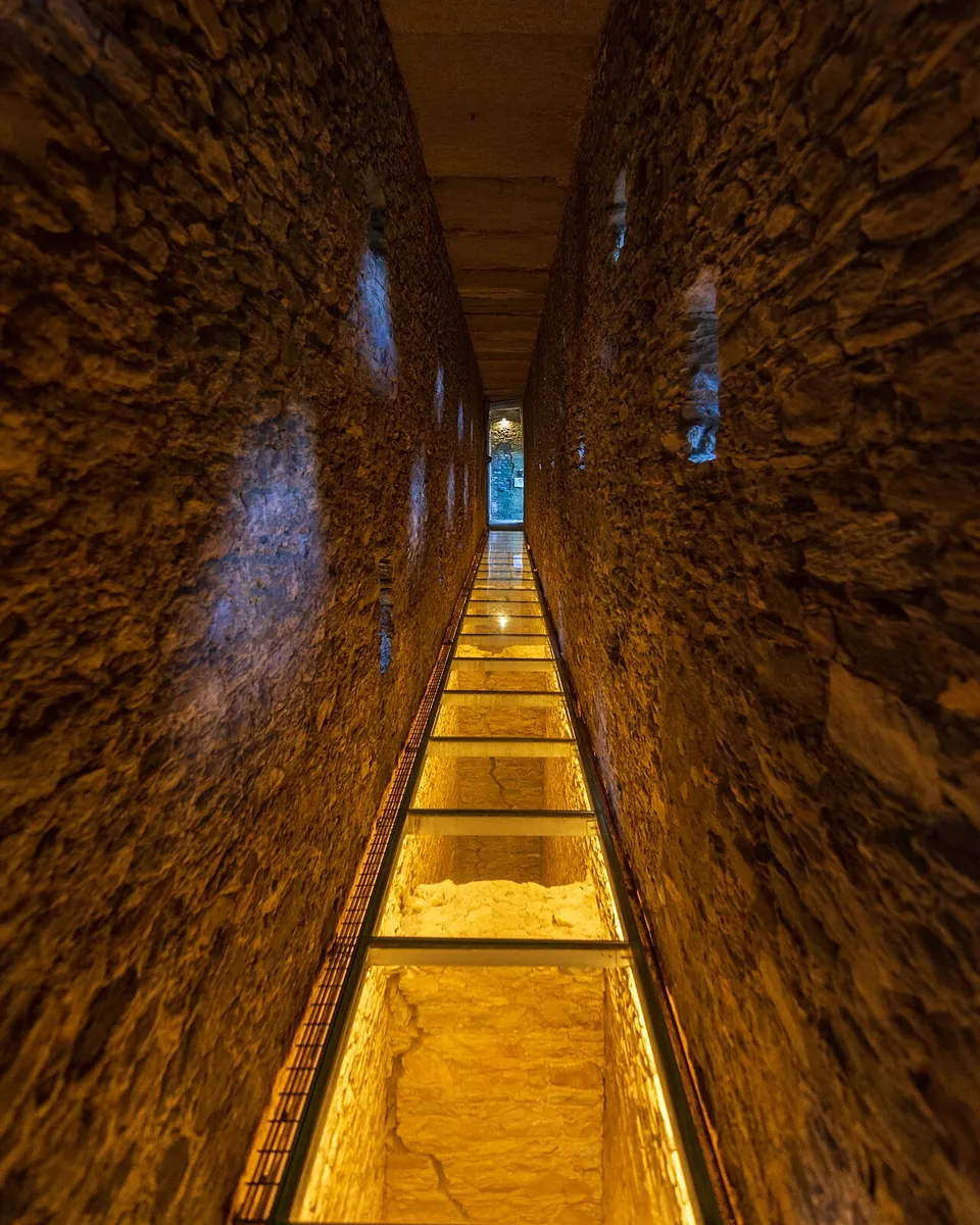 A long, narrow passage between rough stone walls, lit warm yellow, with small openings in the walls and a walkway of glass panels lit from below running into the distance.