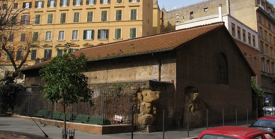 A low brick building with a pitched tiled roof and an arched window, inside a small railed garden with benches at a street corner, overlooked by tall apartment blocks; two weathered stone blocks stand at its front corner.