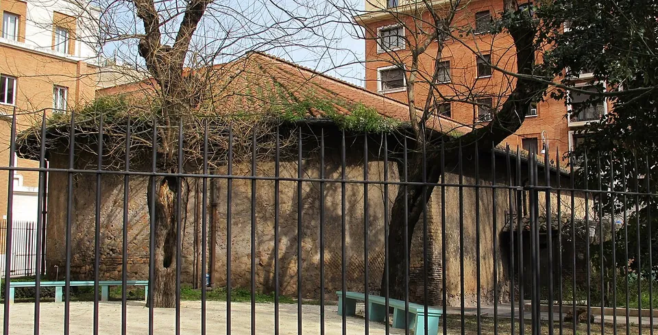 A curved wall of rough masonry with plants growing along its top, seen through tall iron railings across a paved garden with benches and bare trees.