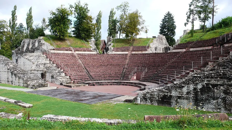 A semicircle of stone seating rising in tiers above a level orchestra floor, framed by rough masonry walls, with trees on the slope above.