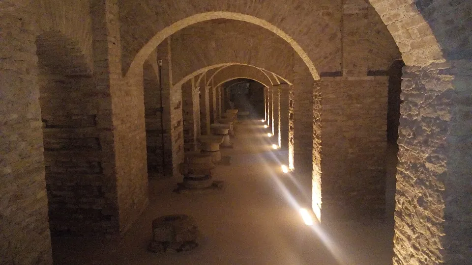 A long underground corridor of brick arches lit from below, with round stone drums set along the floor.