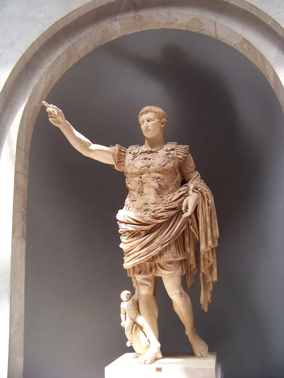 A white marble statue of a man in an ornately carved breastplate and a cloak draped over one arm, his right arm raised, a small figure at his right leg, standing in an arched niche.