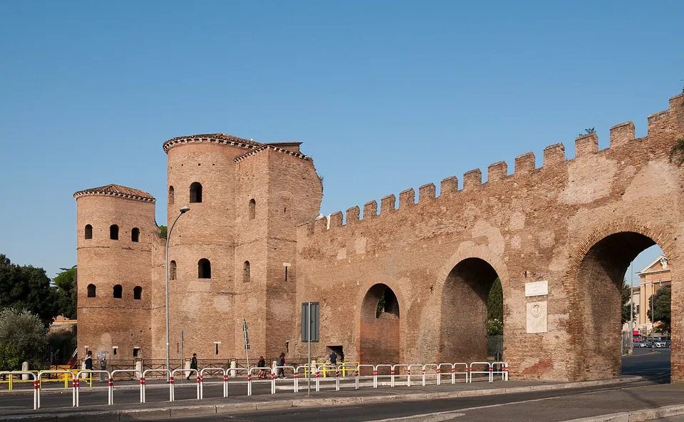 A stretch of battlemented brick city wall with two tall round towers at the left and large arched openings through the wall at the right, with a road and railings in front.