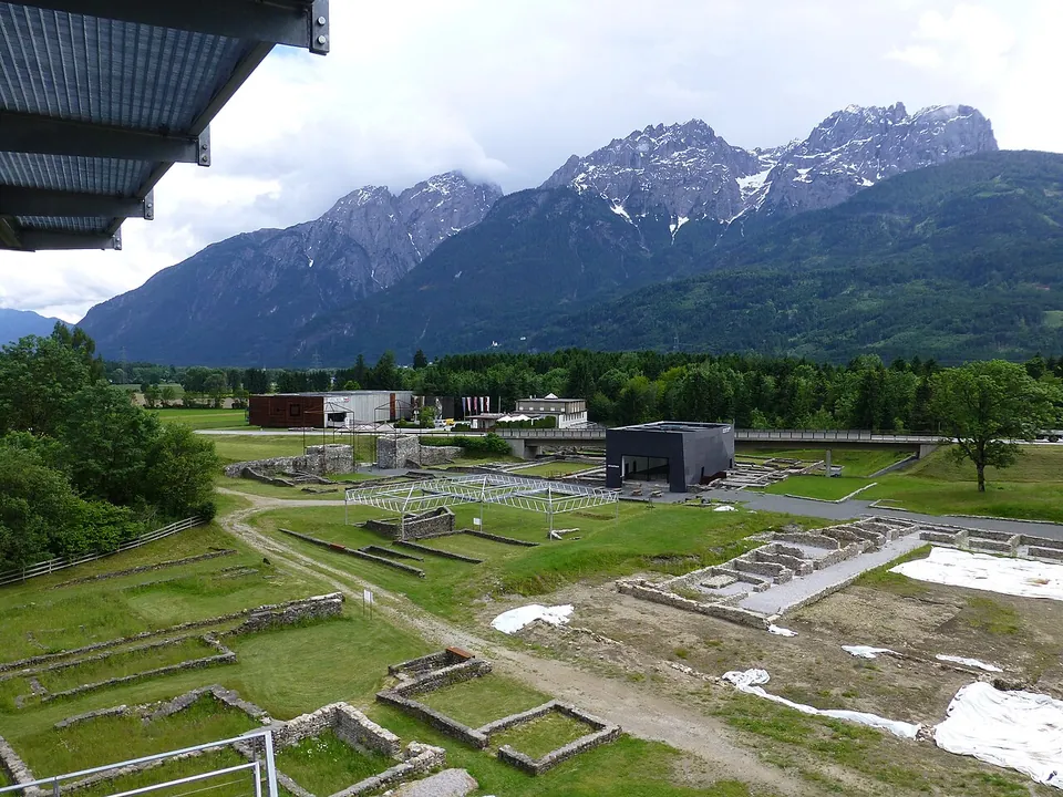 A view down over grassy excavations with low wall outlines and paths, a dark museum building beyond a road, and jagged mountains with patches of snow on the horizon.