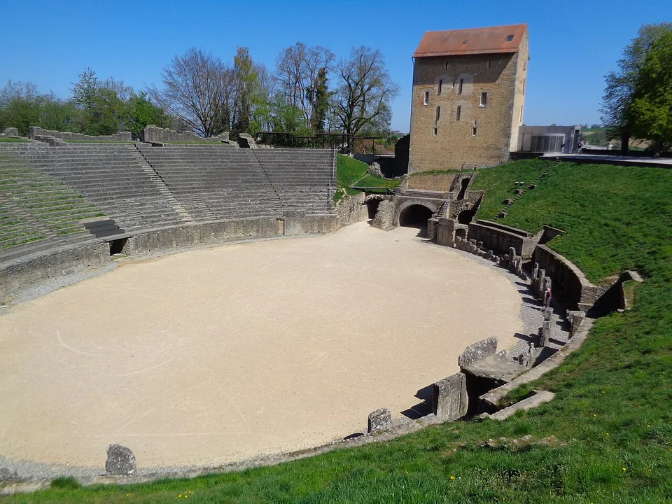 A sanded oval arena ringed by a low stone wall, with tiers of seating rising on the left and a square stone tower with a red tile roof standing over an arched entrance on the far side.