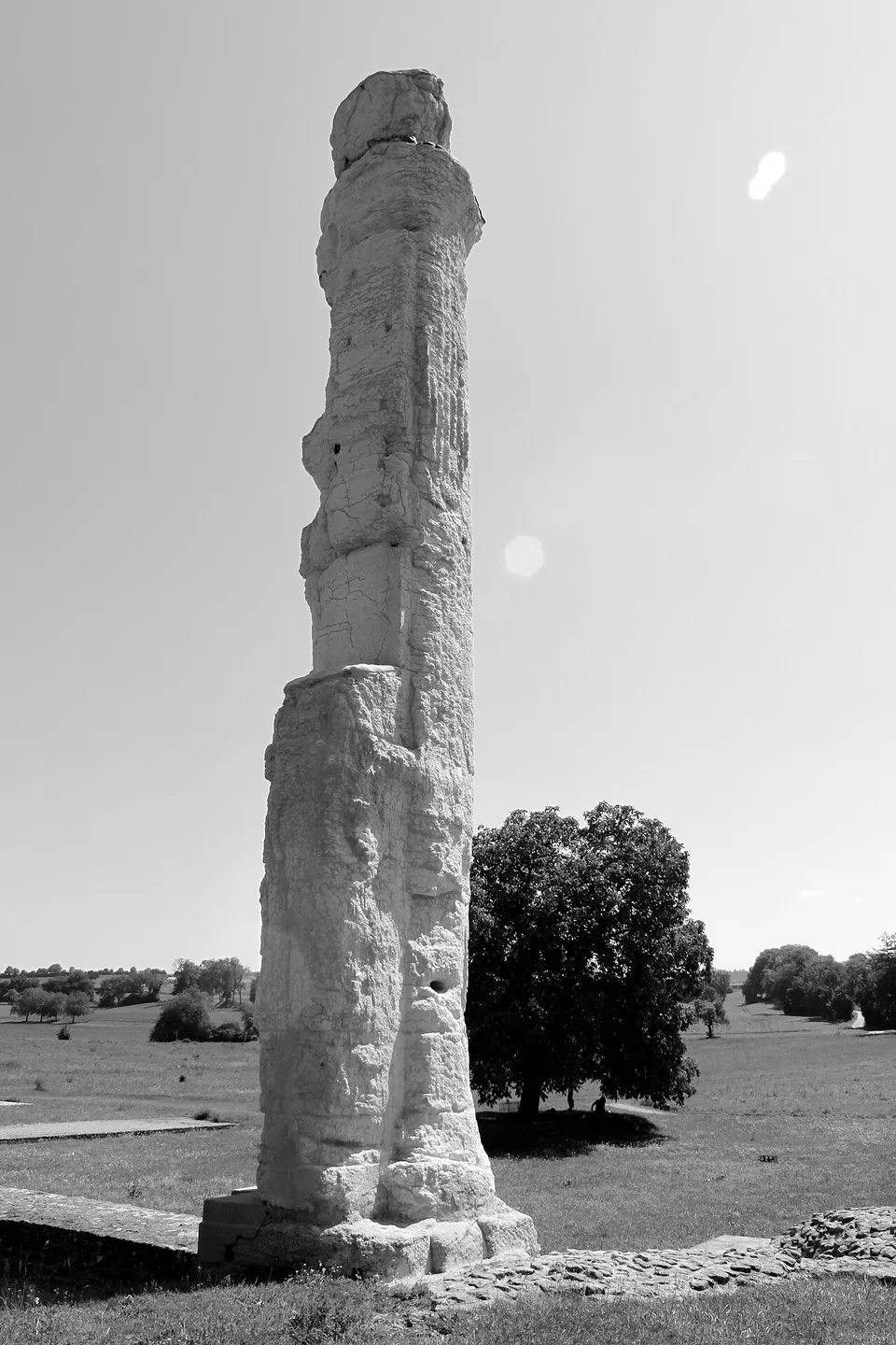 A single tall weathered stone column on a block base standing alone in open grassland, photographed in black and white.