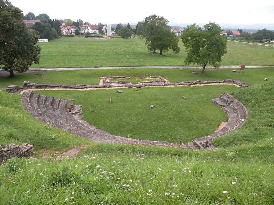 A grassy hollow in a hillside lined with curved rows of low stone seating, the outline of the stage building marked by low walls beyond, with fields, a road and houses behind.