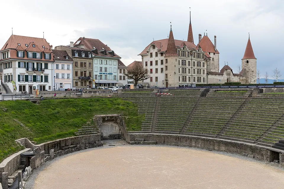 The oval arena of the amphitheatre with grassy and stone tiers of seating, and behind its rim the houses of Avenches and the white château with its pointed towers.