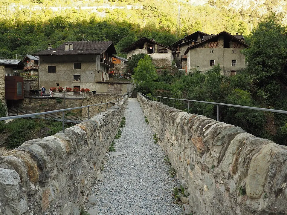 A gravel path between two low stone parapets runs across the top of the bridge towards the stone houses of the hamlet, with metal handrails on either side and wooded slopes behind.