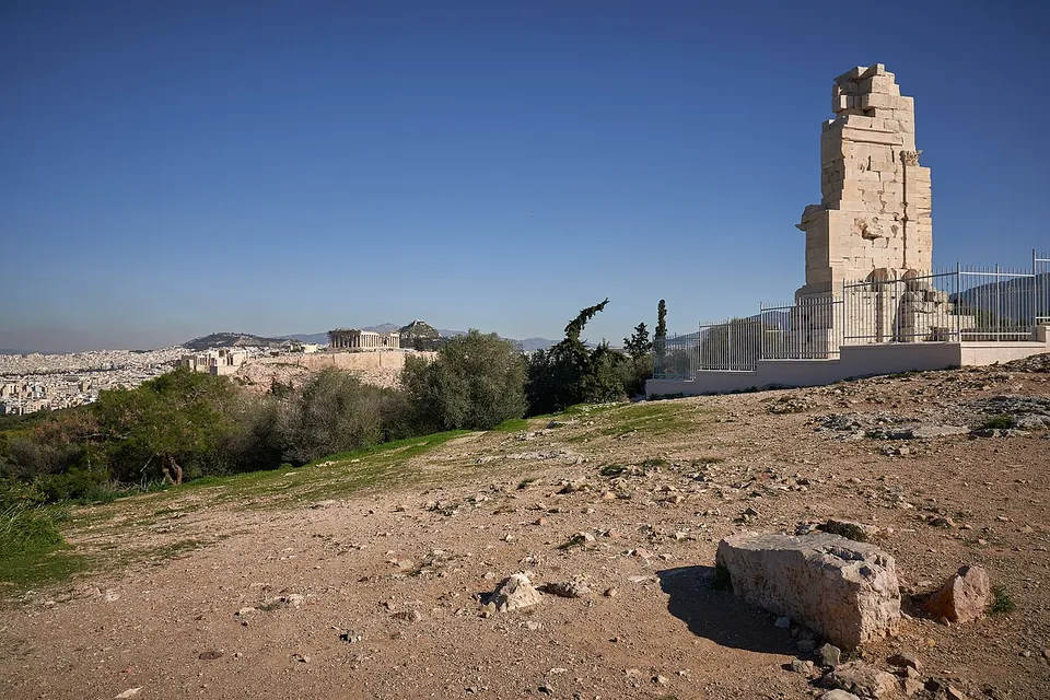 The plain back of a tall stone monument behind a railing on a bare rocky hilltop, with the Acropolis and its temples low on the horizon to the left and the city spread out beyond.
