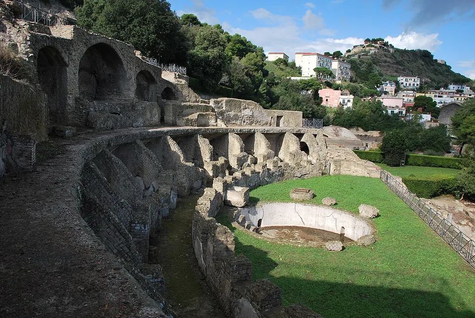 Ruined baths built in terraces into a hillside, a curving row of vaulted stone rooms above a long corridor, with a lawn and a round basin on the lower level and houses on the hill behind.