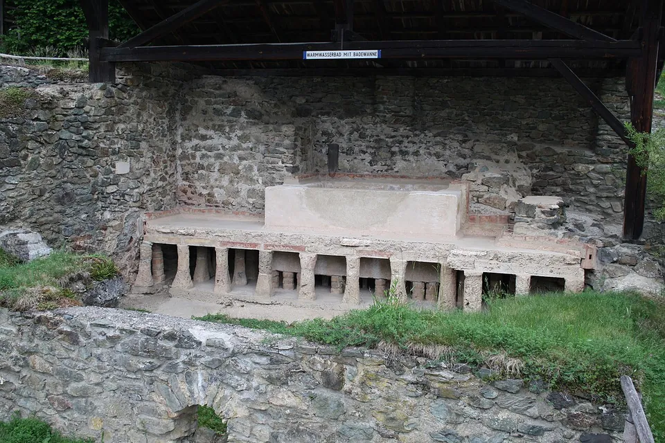 A raised stone bathing tub on short stone pillars, set against rubble walls under a timber roof, with a sign above it.