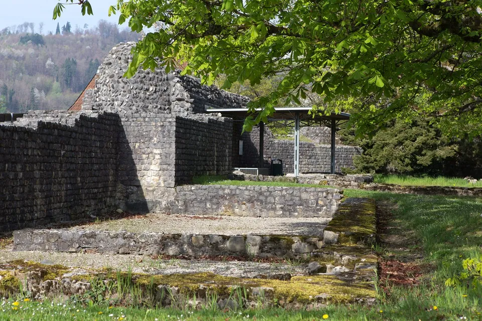 Low conserved walls of rough stone rising in steps towards a taller ruined block of masonry, with a flat roof on slender posts behind and trees in spring leaf.