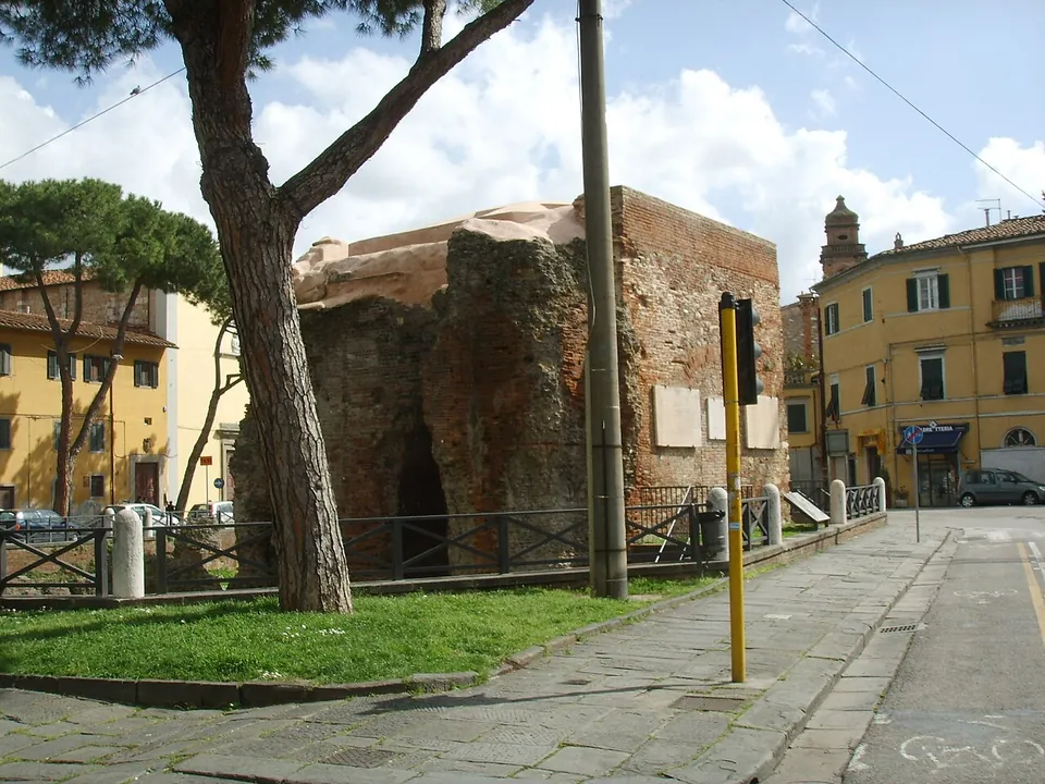 A paved pavement running along a street past a pine tree and metal railings, with the brick and rubble ruin behind the railings on the left and yellow town houses ahead.