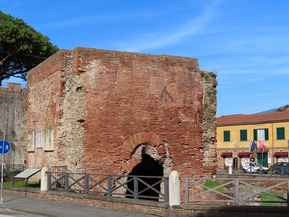 A square block of red brick ruin with a dark arched opening at its base, standing inside a wooden-railed enclosure beside a paved street, an information board at the railing, an old brick wall behind it on the left and yellow-fronted houses on the right.