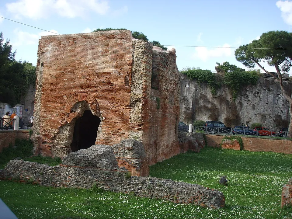 The brick block of the ruin seen across mown grass, with a low stretch of rubble wall running in front of it and a tall weathered wall behind on the right.