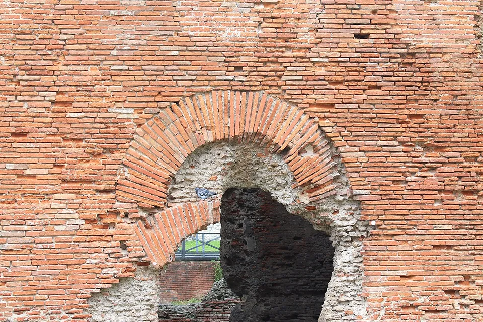 Close view of a wall faced with thin red bricks in regular courses, an arch of bricks set on edge over an opening, and rough rubble core showing where the facing has gone.