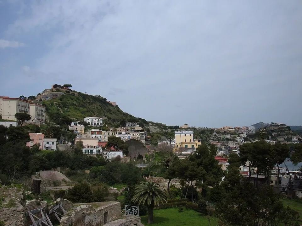 Low ruined walls and vaults among trees and palms in the foreground, with the houses of Baia climbing a green hill behind them.