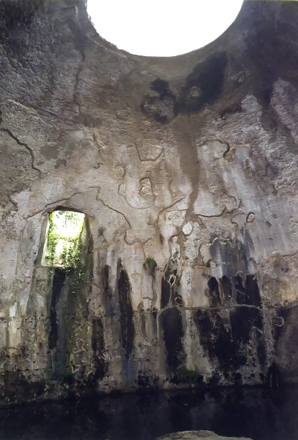 Looking up inside a dark, weathered dome to the round opening at its crown, with a small window in the wall and still water at the bottom.