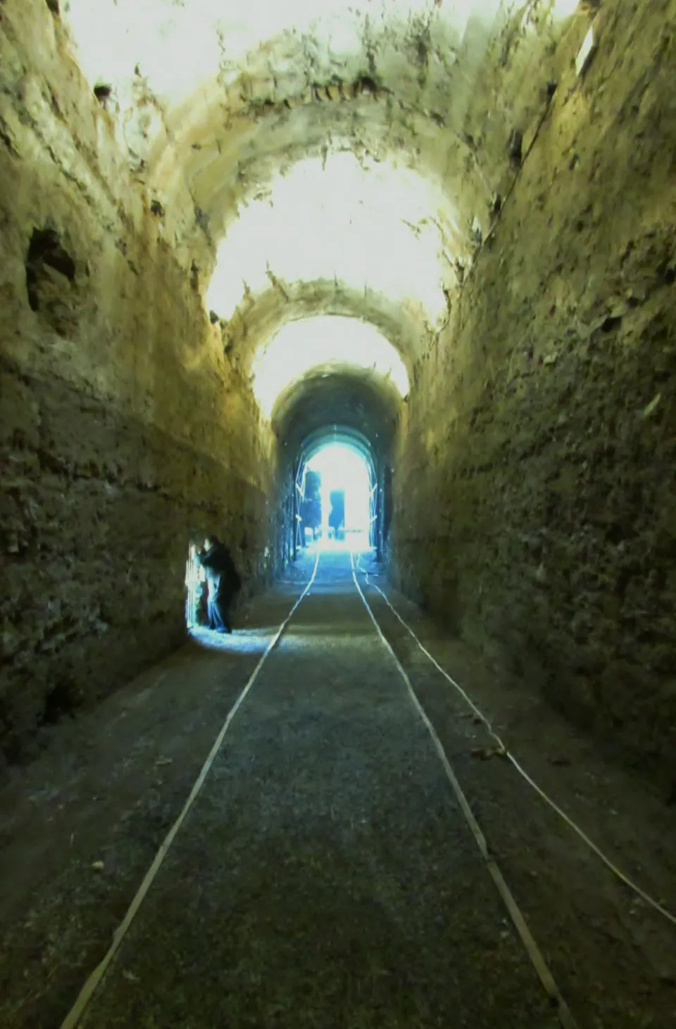 A long, narrow barrel-vaulted corridor with rough concrete walls, lit at the far end, a figure standing in the light.