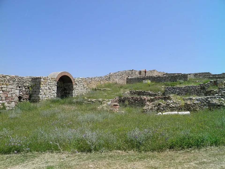A long stone wall with a brick arch running across a grassy slope, with lower excavated walls in front under a clear blue sky.