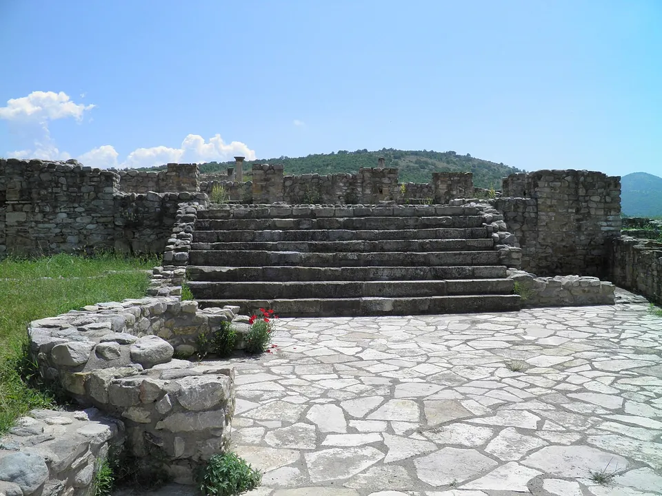 A flight of broad stone steps rising from an irregular stone paving between two stretches of rubble wall, with a wooded hill behind.