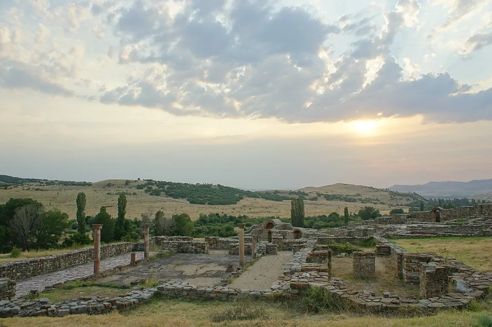 Excavated stone walls and a few standing columns of a large church complex in dry grassland at sunset, with hills and scattered trees behind.