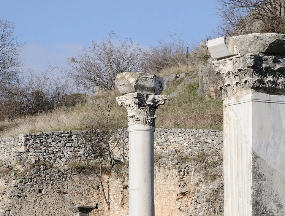 Two white marble columns topped with carved capitals and blocks of stone, against a stone wall and a scrubby slope.