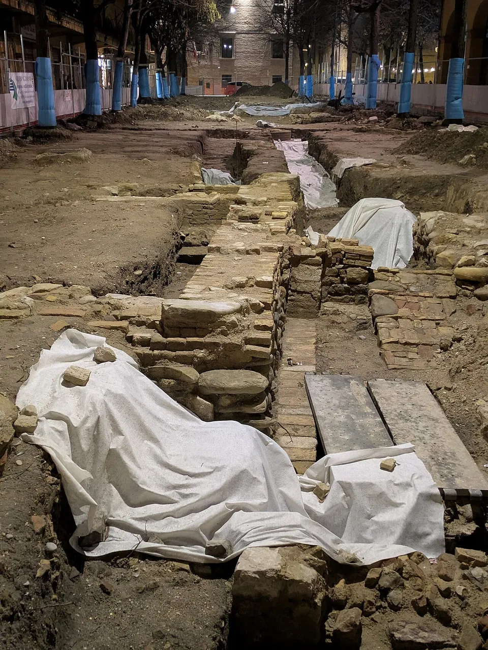 An excavation trench running down the middle of a fenced town square at night, with a line of stone and brick masonry partly covered by white sheeting and planks, trees wrapped in blue protection and buildings beyond.