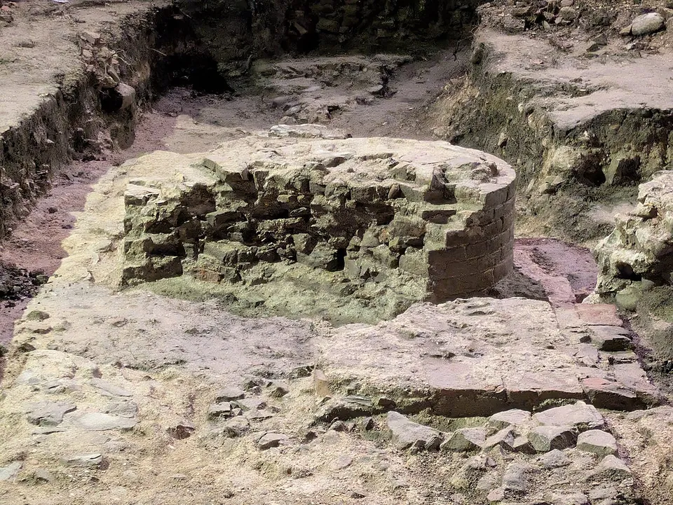 A round stump of rough stone and brick masonry sitting in an excavated surface of compacted earth and stone, with a trench cut behind it.