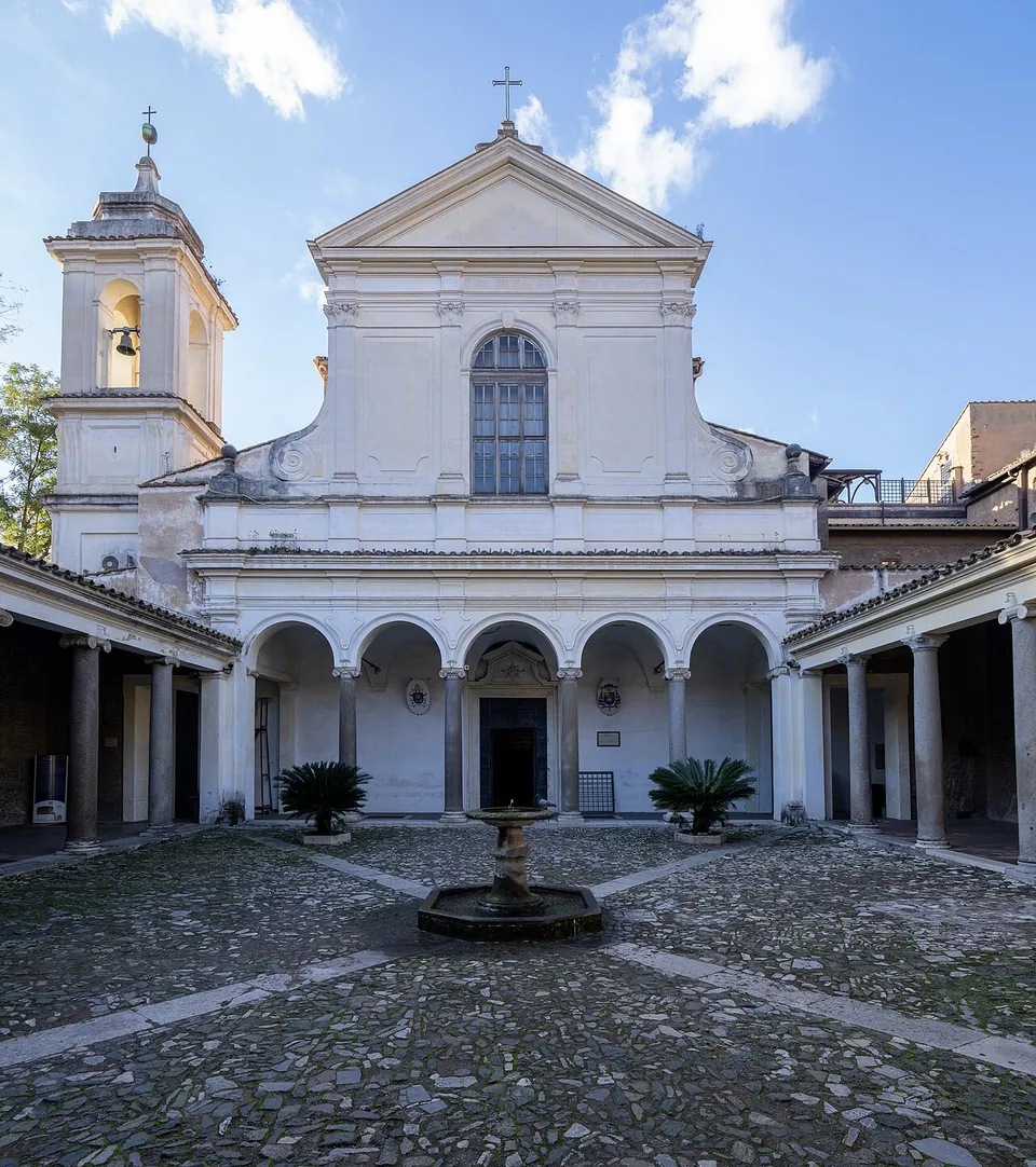 A cobbled courtyard with a small fountain in the middle, ringed by arcades on columns, and behind it a white church facade with a pediment, a large arched window and a bell tower to the left.