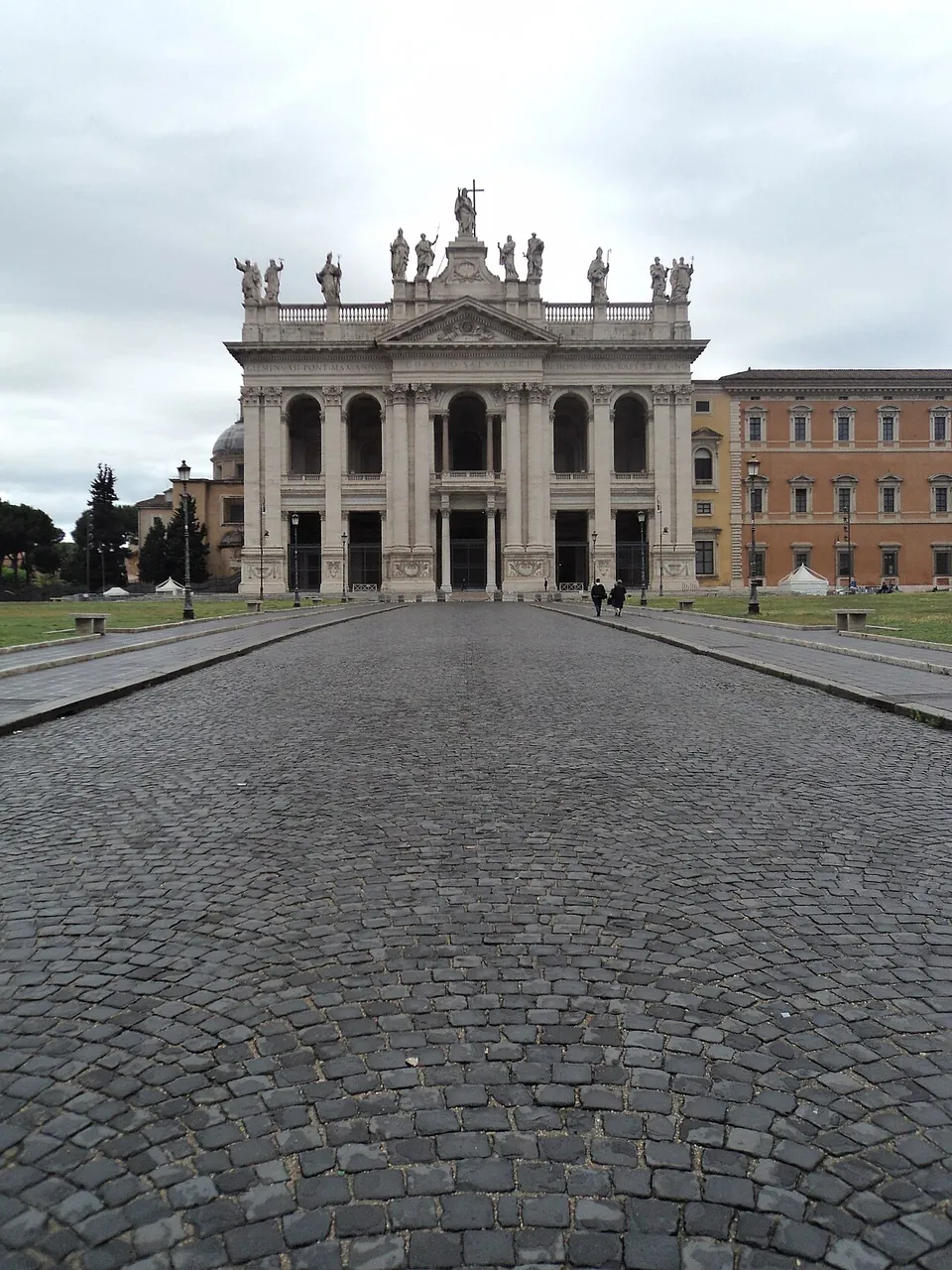 A long cobbled avenue running straight towards a white stone church front with columns, arched openings and a row of statues along its roofline, with the orange palace to its right.