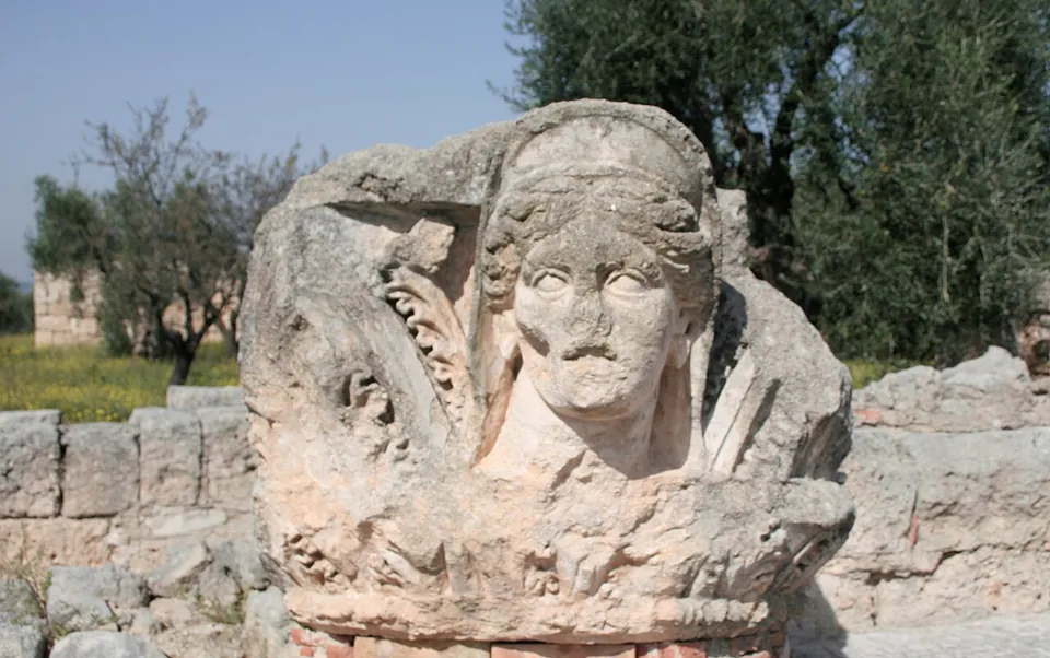 A weathered limestone capital carved with a woman's head framed by leaves, set on a low dry-stone wall among olive trees.