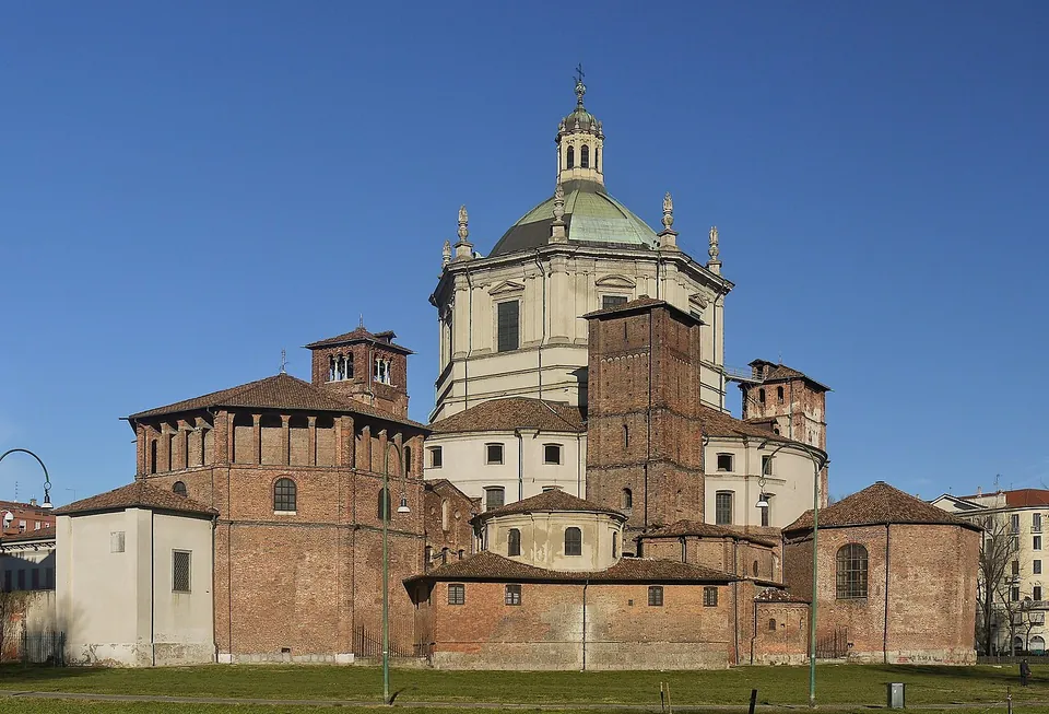 A large brick church complex of several blocks and square towers, crowned by a tall pale octagonal drum with a green dome and lantern, behind a lawn.
