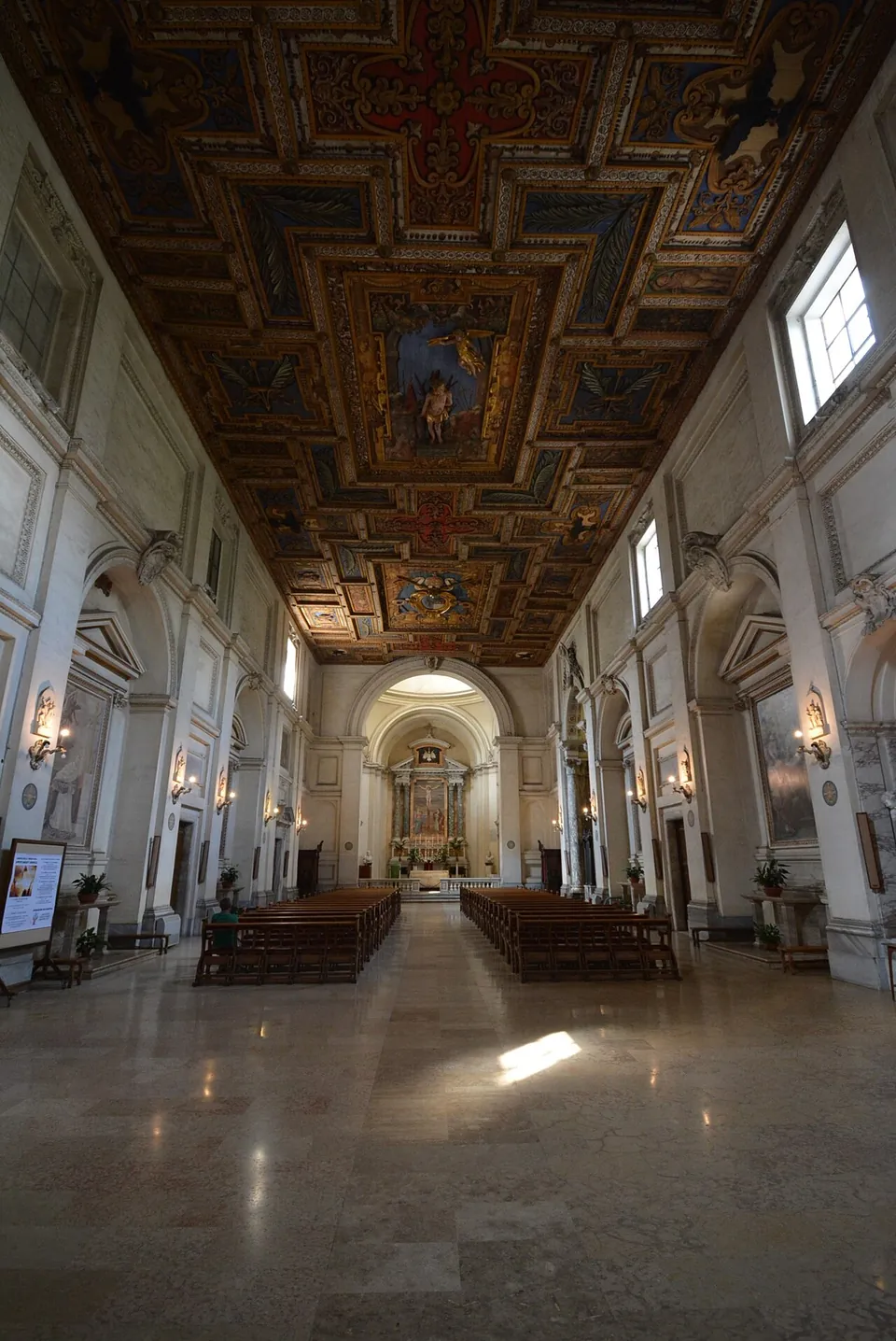 A long single-nave church interior with pale walls, wall lamps and rows of wooden pews under a dark carved and painted wooden ceiling, leading to an altar under an arch.