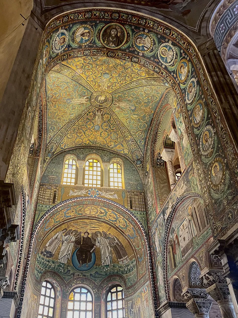 Looking up into the sanctuary. A cross-vault covered in green and gold mosaic rises above three arched windows; arches around it are lined with portrait medallions, and in the half-dome of the apse below, a figure in purple sits on a blue globe between standing figures.